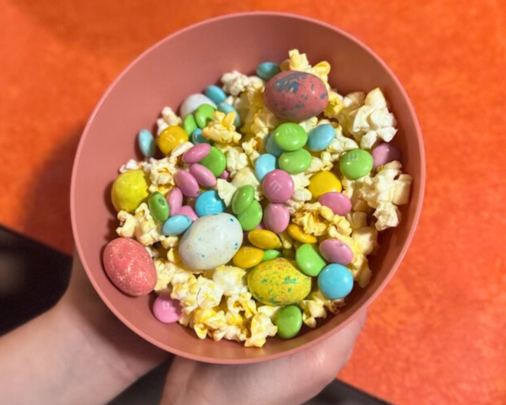 Hand holding a bowl of Easter snack mix with popcorn, pastel candy, and chocolate eggs
